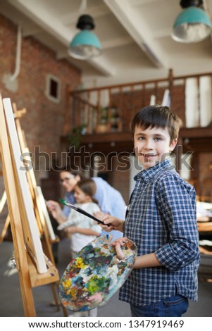 Portrait of smiling boy painting picture on easel in art class and holding palette, copy space
