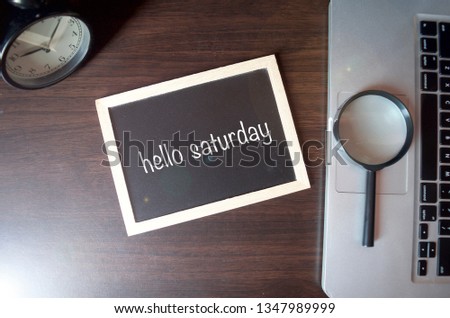 Blackboard writing "hello saturday" on wooden desk background with laptop, magnifying and alarm clock. Conceptual image. Selective focus.