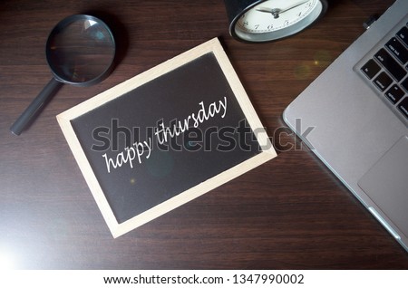 Blackboard writing "happy thursday" on wooden desk background with laptop, magnifying and alarm clock. Conceptual image. Selective focus.