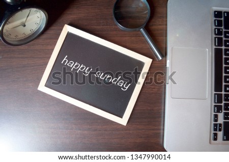 Blackboard writing "happy sunday" on wooden desk background with laptop, magnifying and alarm clock. Conceptual image. Selective focus.