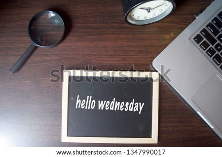 Blackboard writing "hello wednesday" on wooden desk background with laptop, magnifying and alarm clock. Conceptual image. Selective focus.