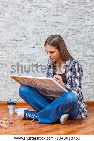 portrait of young teenager brunette girl with long hair sitting on floor and drawing picture on gray wall background with copy space