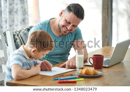 Young dad with green highlighter helping his little son draw a picture on paper while both sitting by table