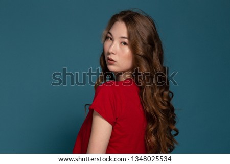 horizontal portrait of a beautiful Asian girl in a red T-shirt and jeans looking into the camera and straightens her long wavy hair on a blue background