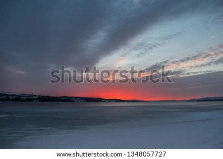 Magnificent and bloody red sunrise over the partly frozen Mjøsa lake in Gjøvik, Norway during winter. Dramatic sky with mountains and forest.