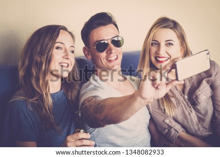Two girls and a young cute man together taking a selfie picture with modern cellular phone to share on social media networks account - technology for cheerful people in friendship sitting on a sofa