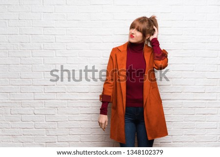 Woman with coat over white brick wall having doubts while scratching head