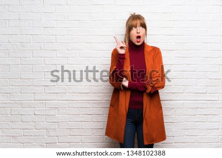 Woman with coat over white brick wall thinking an idea pointing the finger up