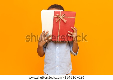 Horizontal image of unrecognizable dark skinned little boy wearing shirt covering face with white and red box with birthday gift, candies or sweets inside, posing at blank yellow studio wall