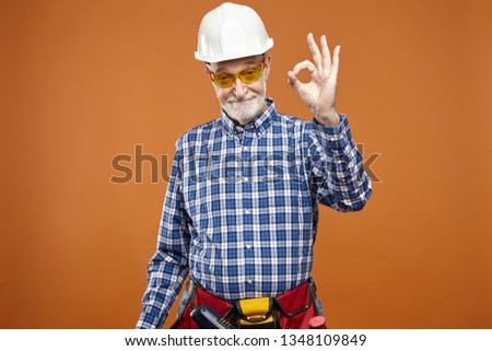 Everything is under control. Portrait of eldelry mature Caucasian handyman with thick beard wearing safety helmet and tool belt, making ok gesture, saying that he will fix broken leaky faucet