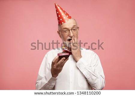 Isolated image of handsome emotional unshaven Caucasian male wearing glasses, white shirt, bow tie and holiday hat covering his opened mouth, having amazed look, holding birthday cupcake with candle