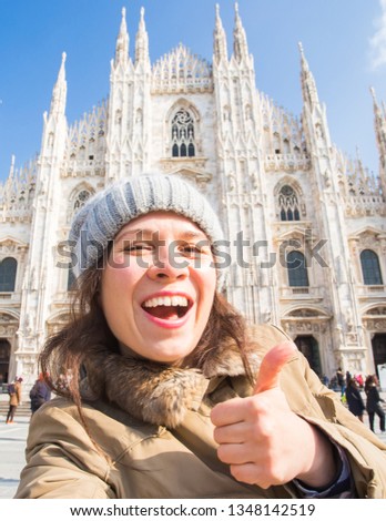 Young happy woman touris making selfie photo and showing thumb up in front of the famous Duomo cathedral in Milan.