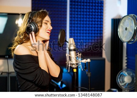 Half-length portrait of a singer with earphones wearing black dress on the blue studio background. Singing woman with studio microphone. Concept of music
