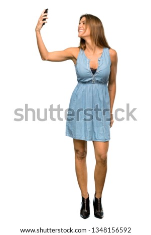 A full-length shot of a Blonde woman with jean dress making a selfie over isolated white background