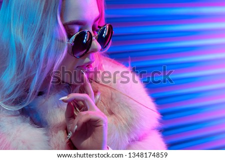 Beautiful teen girl in stylish glasses fur on neon wall background, blue pink purple street lights fashion trendy teenager hipster young woman face retrowave synthwave glowing night life city style
