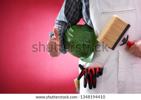 Caucasian house painter worker in white work overalls, with helmet and brush making ok sign with thumb up. Construction industry. Work safety. Red background.