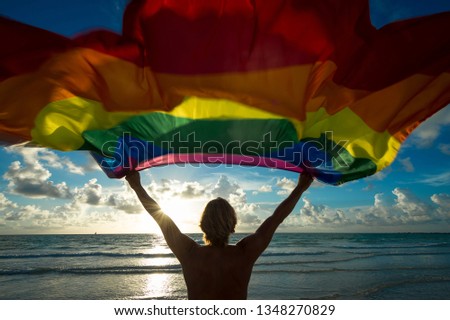 Colorful silhouette of a man with blond hair holding a gay pride rainbow flag blowing in the wind on a tropical beach with golden sun