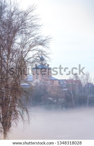 Peaceful spring misty landscape with fog at sunrise after rain with rainbow on the dirty river Volga, with orthodox church on the hill in the background.