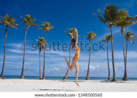 happy woman in a yellow bathing suit running along the beach with coconut trees and jumping up    