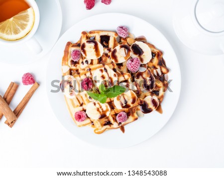 Belgian curd waffles with raspberries, banana, chocolate syrup.  Breakfast with tea on white background, top view