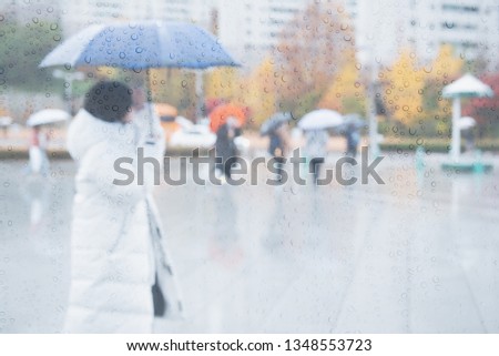 Walking in the rain. Raindrops on glass wall with blurred woman and crowd people with umbrella walking on pedestrian, raining with colorful autumn leaves background, can be used for weather forecast