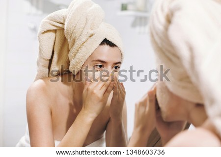 Young happy woman in towel making facial massage with  organic face scrub and looking at mirror in stylish bathroom. Girl applying scrub cream, peeling and cleaning skin. Skin Care Royalty-Free Stock Photo #1348603736