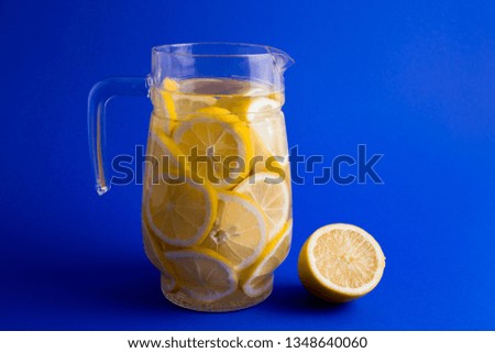 Transparent glass jar full with lemon slices and fresh, refreshing lemonade. Isolated on a bold, clean, blue background. 