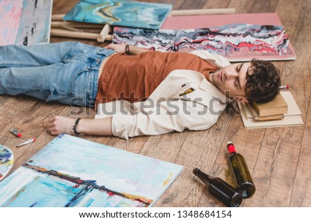exhausted artist lying on wooden floor, surrounded with paintings, draw utensils and empty bottles