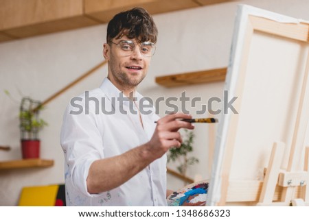 selective focus of smiling artist in white shirt and glasses painting on canvas and looking at camera