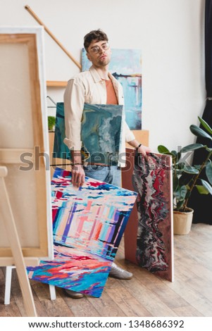 serious artist in eyeglasses holding colorful paintings and looking at camera