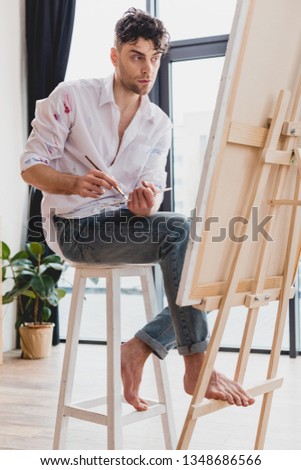 handsome artist in white shirt and blue jeans mixing paints on palette while sitting at easel in gallery