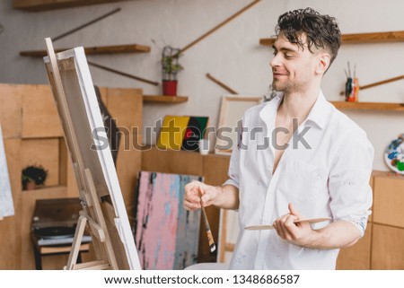 selective focus of smiling artist in white shirt and blue jeans looking at canvas