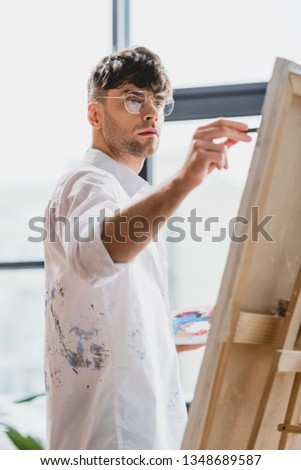 selective focus of handsome artist in glasses painting on canvas