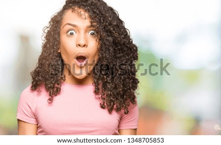 Young beautiful woman with curly hair wearing pink t-shirt afraid and shocked with surprise expression, fear and excited face.
