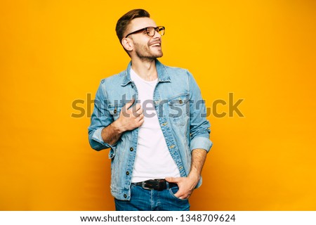 Holding the moment. Fair-faced man in the denim outfit which consist of a jacket and jeans mixed with bright white t-shirt and fashionable glasses in front of yellowish wall.