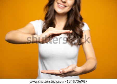 Thin and tall girl is showing a right-angled shape with her hands while staying in front of goldish background wearing white basic t-shirt.