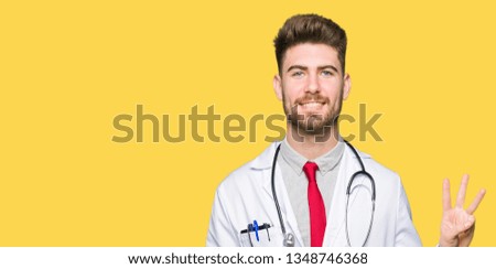 Young handsome doctor man wearing medical coat showing and pointing up with fingers number three while smiling confident and happy.