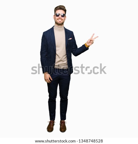 Young handsome elegant man wearing sunglasses smiling with happy face winking at the camera doing victory sign. Number two.