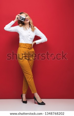 Full length of a beautiful young blonde woman standing over red background, holding photo camera