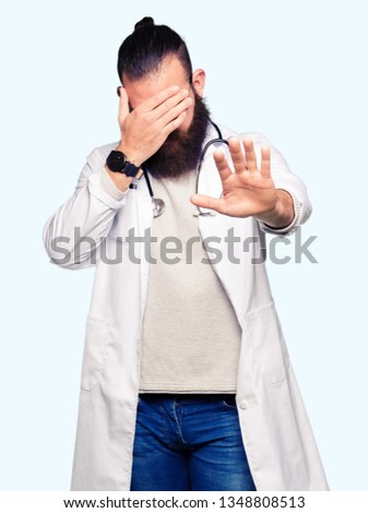 Young blond doctor man with beard wearing medical coat covering eyes with hands and doing stop gesture with sad and fear expression. Embarrassed and negative concept.