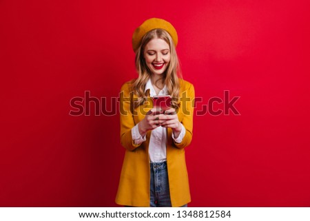 Blithesome stylish woman holding smartphone isolated on red background. Studio shot of curious french girl using phone.