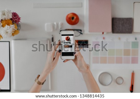 Elegant female hands with black manicure hold iPhone with picture of workplace on screen. Laptop, flowers, apple, documents are on white background