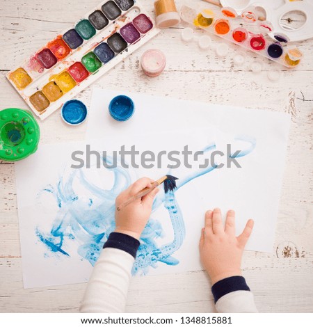 Top view on the hands of the kid with a brush painting on a sheet of paper. Workspace for artwork with watercolors, glass of water, palette and brushes. Art tools on a white wooden table.
