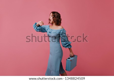 Beautiful european woman in blue outfit looking at wristwatch. Indoor portrait of emotional dark-haired lady with little valise.