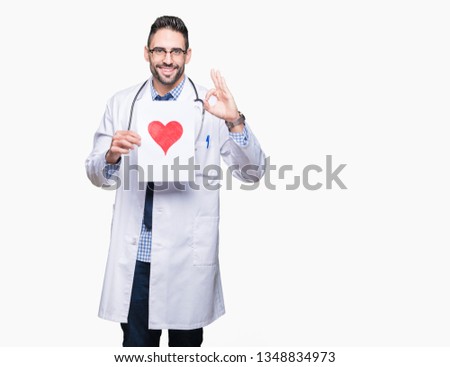 Handsome young doctor man holding paper with red heart over isolated background doing ok sign with fingers, excellent symbol