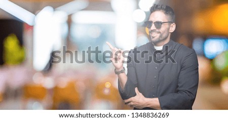 Young Christian priest wearing sunglasses over isolated background with a big smile on face, pointing with hand and finger to the side looking at the camera.