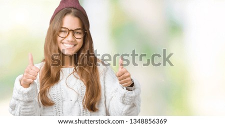 Young beautiful brunette hipster woman wearing glasses and winter hat over isolated background success sign doing positive gesture with hand, thumbs up smiling and happy. Looking at the camera
