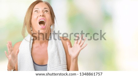 Beautiful middle age woman wearing sport clothes and a towel over isolated background crazy and mad shouting and yelling with aggressive expression and arms raised. Frustration concept.