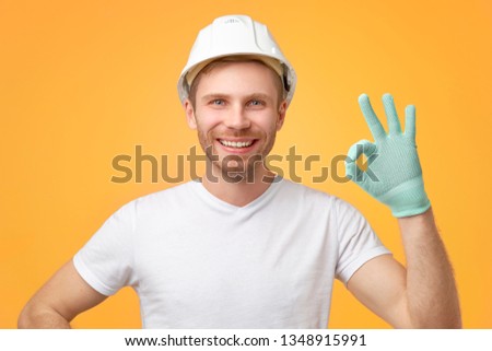 Pleased confident European man with broad smile, uniform, shows okay gesture, dressed in t-shirt and construction helmet. Isolated over white background. 