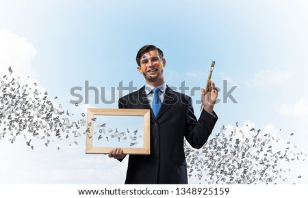 Portrait of young and successful businessman in black suit holding picture frame and paintbrush in hands while standing with flying letters against blue cloudy skyscape view on background.